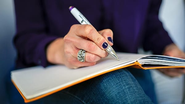 A person writing in an open notebook with a pen at a desk beside scattered papers
