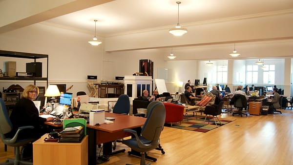 An open-plan office interior with desks, shelves and filing under daylight