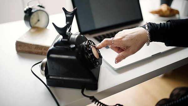 A hand dialing a rotary telephone standing on a desk beside a laptop