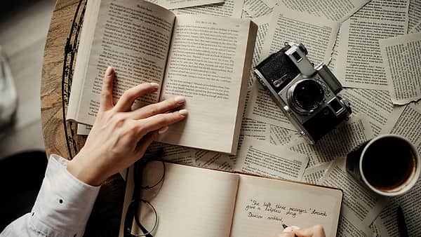 Hands holding an open book while a person writes in a notebook surrounded by papers